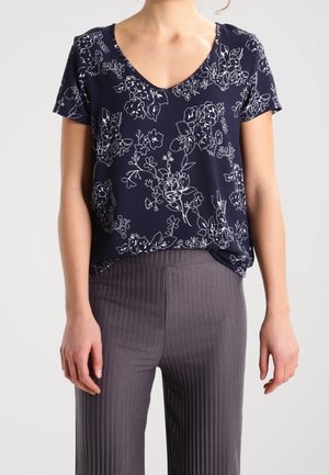 Femme portant un haut bleu marine à manches courtes avec un col en V, orné de contours floraux blancs, et un pantalon taille haute à rayures grises.