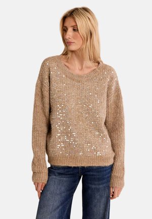 Donna con lunghi capelli biondi che indossa un maglione lavorato a maglia beige con paillettes e jeans blu, che guarda di lato su uno sfondo semplice.