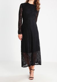 Kvinna iklädd en lång svart spetsklänning med långa ärmar och öppna svarta klackar med ankelrem mot en enfärgad vit bakgrund.