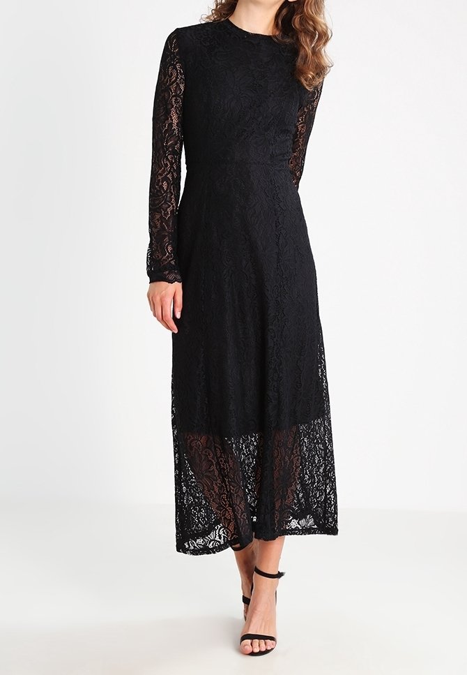 Kvinna iklädd en lång svart spetsklänning med långa ärmar och öppna svarta klackar med ankelrem mot en enfärgad vit bakgrund.