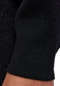 Schwarzer gerippter Strickstoff mit metallischen silbernen Sprenkeln. Nahaufnahme des Ärmelbundes, der eine strukturierte Oberfläche und ein elastisches Band zeigt.