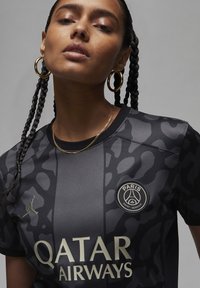 Nainen, jolla on letitetyt hiukset, pukeutuneena kultaisiin rengaskorvakoruihin ja mustaan camotyyppiseen Paris Saint-Germain -paitaan, jossa on Qatar Airways -logo.