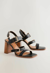Sandali neri con tacco alto, caratterizzati da tre fasce larghe e fibbie in tonalità oro. Una punta quadrata e un tacco a blocco in legno texturizzato completano il design.