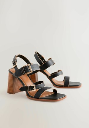 Zwarte hoge sandalen met hakken, voorzien van drie brede banden en goudkleurige gespen. Een vierkante neus en een houten blokhak met textuur maken het ontwerp compleet.