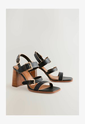 Zwarte hoge sandalen met hakken, voorzien van drie brede banden en goudkleurige gespen. Een vierkante neus en een houten blokhak met textuur maken het ontwerp compleet.
