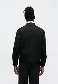 Čierna rifľová bunda s textúrovaným vyšívaným dizajnom koňa na chrbte. Má golier a manžety na gombíky. Nosená s tmavými džínsami.