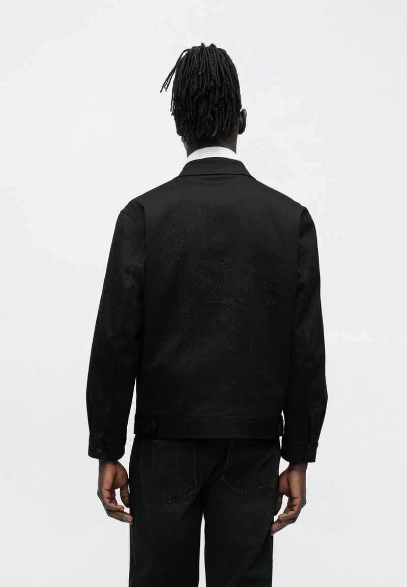 Schwarze Jeansjacke mit einem strukturierten, bestickten Pferdemotiv auf der Rückseite. Verfügt über einen Kragen und knöpfbare Manschetten. Getragen mit dunkler Jeans.