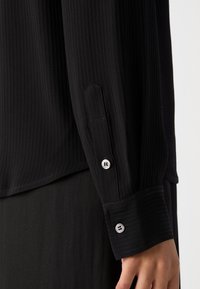 Chemise noire à manches longues côtelée avec un poignet boutonné décoré de boutons métalliques. Le tissu a un aspect texturé et un design épuré.