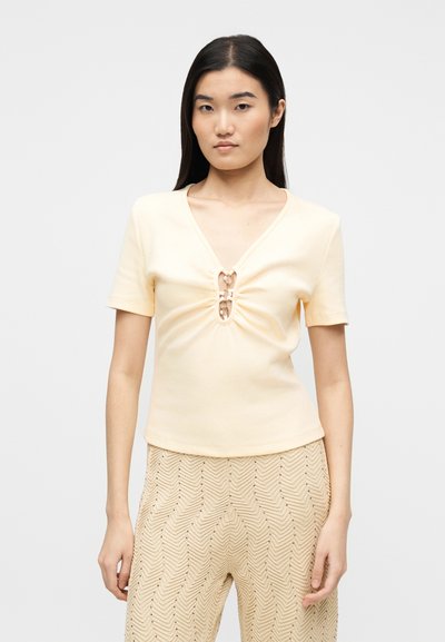 Νεαρή γυναίκα με ίσια σκούρα μαλλιά, φοράει κοντομάνικη ανοιχτόκίτρινη μπλούζα με δύο μεταλλικά κλιπ μπροστά και μπεζ παντελόνι με σχέδια.