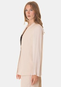 Blazer beige con maniche lunghe plissettate, scollatura a V e vestibilità sagomata. Realizzato in tessuto morbido, con dettagli di cucitura sottile.