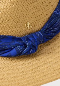Ψάθινο καπέλο με μπλε φουλάρι με σχέδια γύρω από τη βάση. Διαθέτει χρυσό έμβλημα "RL" στην κορυφή. Φυσική υφή και ανοιχτό καφέ χρώμα.