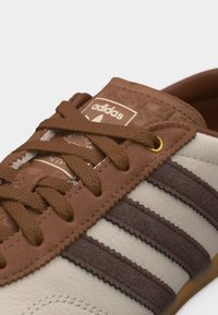 Κοντινό πλάνο καφέ και μπεζ αθλητικού παπουτσιού Adidas με καφέ κορδόνια, τρεις καφέ ρίγες και λογότυπο Adidas στη γλώσσα.