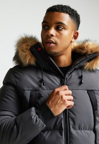 Hall gray puffer jope, mille on kaunistatud karva voodriga kapuutsiga, punaste aktsentidega, tõmbluku sulgemise ja elastsete käistega. Sujuv tekstuur ja keha külge toetav lõige.