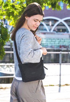 Borsa a tracolla nera con una texture morbida, forma rettangolare e chiusura a patta. Presenta un logo discreto sul davanti. Indossata sopra un maglione grigio.
