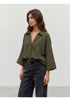 Femme aux longs cheveux bruns portant un chemisier ample vert olive et un jean noir délavé taille haute avec une ceinture noire, debout devant un fond uni.