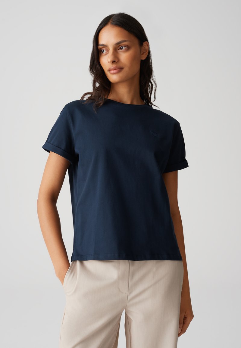 T-shirt in cotone blu navy con maniche arrotolate e colletto a giro; dettaglio logo discreto sul petto sinistro; abbinato a pantaloni beige chiaro.