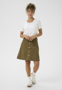 Groene corduroy a-lijn rok met een vooroor zijn knoopsluiting en zakken, gecombineerd met een witte top met gegolfde randen en korte mouwen, en casual sneakers.