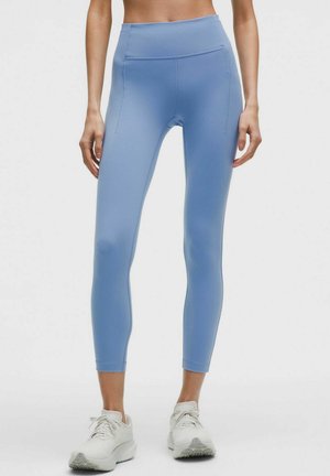 Kvinde iført lyseblå, højtaljede træningsleggings med sidelommer og hvide sneakers, stående mod en ensfarvet baggrund.