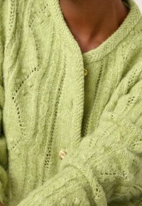 Měkký zelený pletený kardigan s texturovanými vzory a zlatými knoflíky, nošený přes odpovídající zelený svetr.