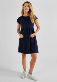Navyblauwe jurk met korte mouwen en een ontspannen pasvorm, met een gerimpelde taille en twee voorzakken. Gecombineerd met witte sneakers.