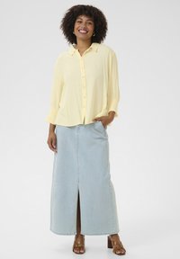 Camicia button-up giallo chiaro con maniche a lunghezza metà abbinata a una gonna maxi in denim azzurro chiaro con uno spacco frontale e sandali marroni con cinturini.
