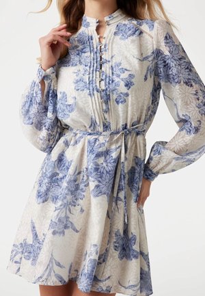 Vrouw draagt een witte jurk met blauw bloemenpatroon, lange doorschijnende mouwen, knopen aan de voorkant en een ceintuur die in een strik is gebonden, tegen een effen achtergrond.