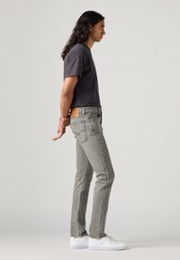 Slim fit szürke farmernadrág bőr folttal, sötét színű pólóval és fehér sportcipővel párosítva. Az öltözet oldalprofilban van megjelenítve.