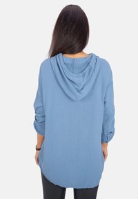 Blauer strukturierter Hoodie mit lässiger Passform, ausgestattet mit Kapuze, langen Ärmeln, die an den Bündchen hochgerollt sind, und einem abgerundeten Saum.