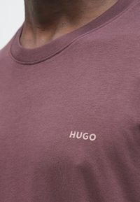 Približen posnetek osebe, ki nosi temno vijolično majico z okroglim izrezom in majhnim svetlim napisom "HUGO" na prsih.