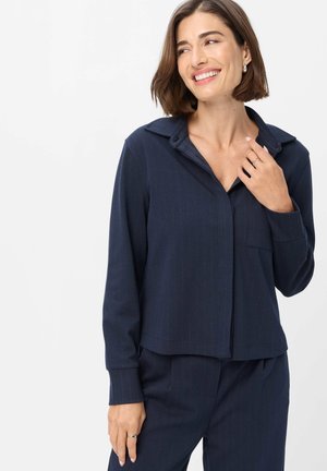 Kvinde med kort brunt hår iført en mørkeblå bluse med lange ærmer og matchende bukser, smilende og kiggende til siden mod en hvid baggrund.