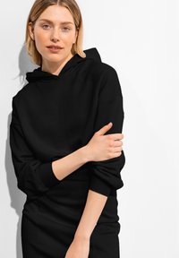 Svart huvtröja med avslappnad passform, ribbade muddar och en mjuk textur, bärs över ett svart plagg, mot en vit bakgrund.