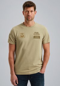 Olivengrøn kortærmet T-shirt i ribbet stof, med broderede lapper på brystet og venstre ærme, samt rund halsudskæring.