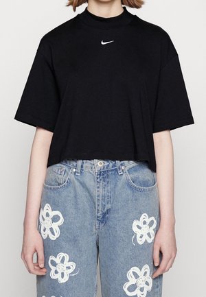 Person trägt ein schwarzes, kurzes Nike-T-Shirt und hochgeschnittene blaue Jeans mit weißen Blumenmustern, steht vor einem schlichten Hintergrund.