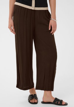 Femme portant un pantalon ample marron foncé, des sandales noires sans bride et un haut noir avec une bordure beige, debout, mains détendues le long du corps.
