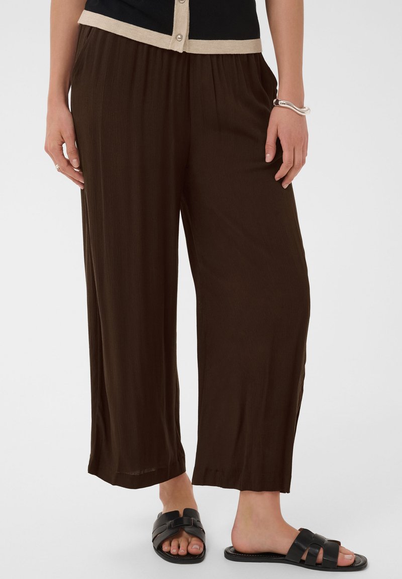 Femme portant un pantalon ample marron foncé, des sandales noires sans bride et un haut noir avec une bordure beige, debout, mains détendues le long du corps.