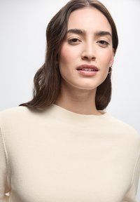 Maglione beige a coste con collo alto e maniche lunghe, caratterizzato da una texture morbida. La modella ha capelli castani di lunghezza media e indossa piccoli orecchini a cerchio.