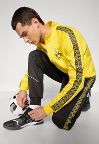 Gul lynlåsjakke med sorte accenter, sorte træningsbukser med lateral design og sorte sneakers. Personen binder snørebånd på en sko ved siden af en fodbold.