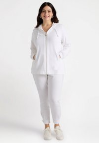 Vit zip-up hoodie med dragsko, som matchas med vita tapered byxor. Mjuk tyg, framfickor och avslappnade sneakers.