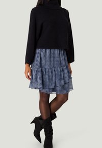 Pull en tricot noir à col roulé associé à une jupe bleue à motifs à deux niveaux, soulignée par des collants opaques noirs et des bottes à talons noires.