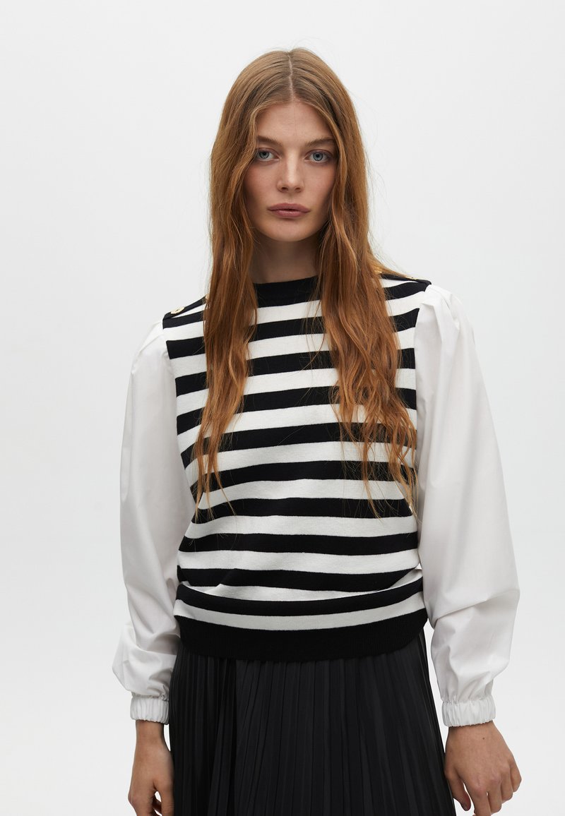 Maglione a righe in bianco e nero con scollatura tonda, tessuto lavorato a maglia e maniche a sbuffo bianche. La gonna plissettata nera completa il look.