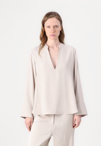 Blusa beige a maniche lunghe con scollatura a V, caratterizzata da una vestibilità rilassata e una texture liscia. Indossata con pantaloni coordinati. Design minimalista.