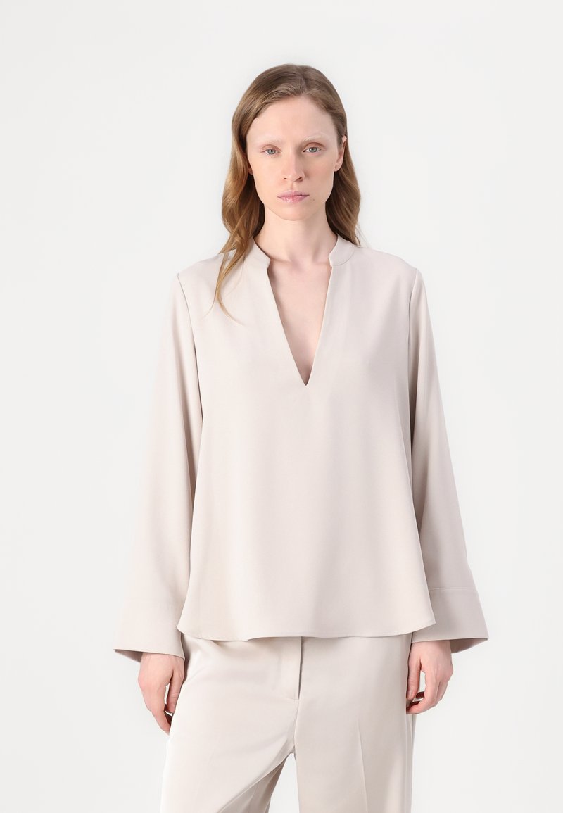 Blusa beige a maniche lunghe con scollatura a V, caratterizzata da una vestibilità rilassata e una texture liscia. Indossata con pantaloni coordinati. Design minimalista.