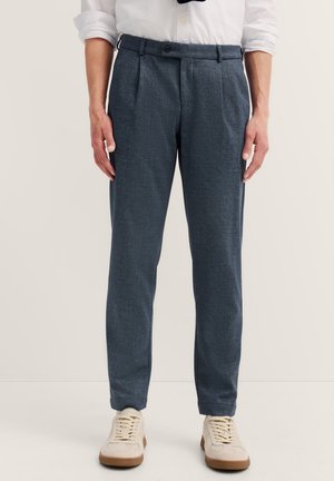 Mand iført mørkegrå skræddersyede bukser, hvide sneakers og en hvid skjorte med opklistrede ærmer, stående mod en ensfarvet baggrund.