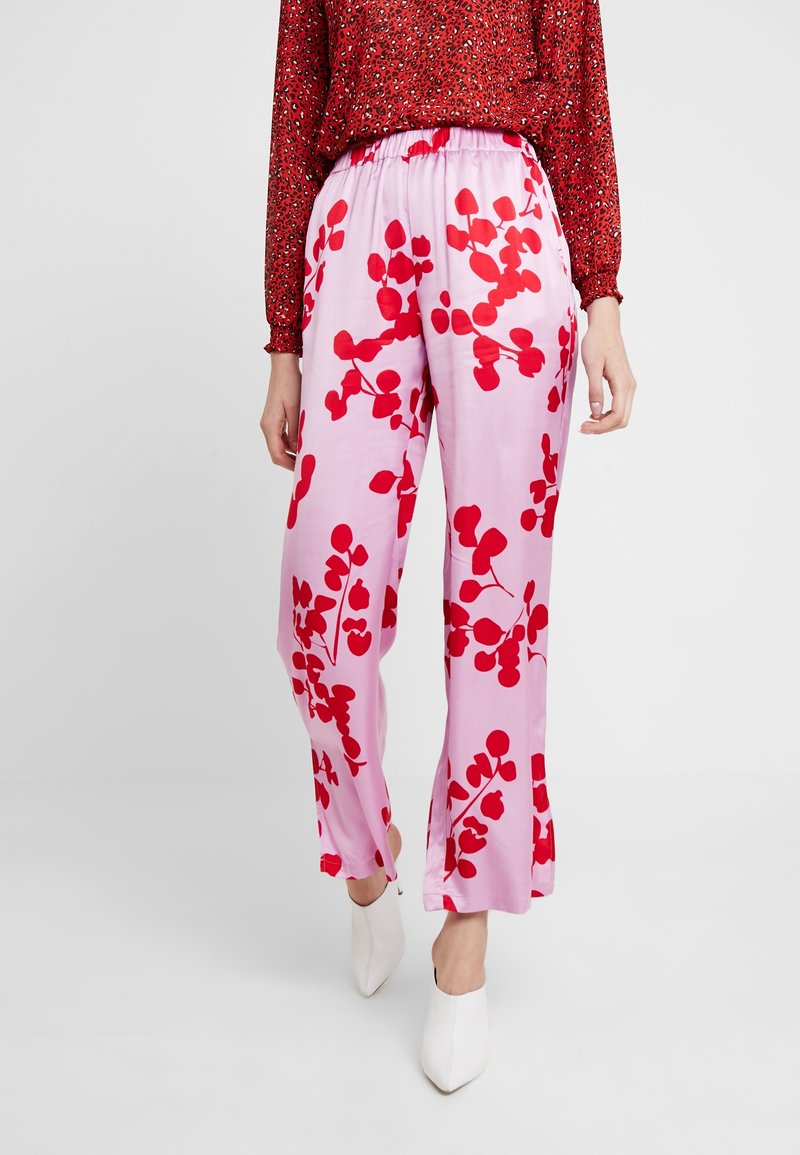 Femme portant un pantalon rose ample avec des imprimés botaniques rouges, associé à un blouse à motif rouge et des chaussures blanches à bout pointu.