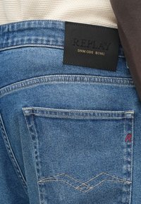 Μπλε τζιν παντελόνι από ντένιμ με μια σκούρα δερμάτινη ετικέτα που γράφει "REPLAY" στη μέση. Διαθέτει ραφές με λεπτομέρειες και ένα μικρό κόκκινο λογότυπο στην πίσω τσέπη.