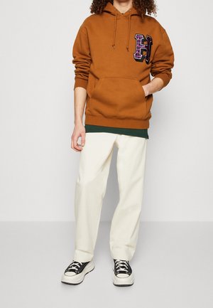 Hnědá bavlněná mikina s kapucí, přední kapsou a velkým fialovým kostkovaným logem "H", doplněná krémovými kalhotami a černými teniskami.