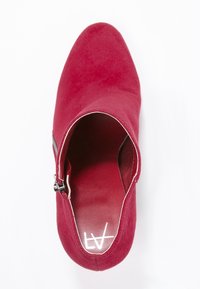 Bottine en daim rouge avec bout pointu, fermeture éclair latérale et extérieur texturé. L'intérieur présente une doublure lisse avec marquage.