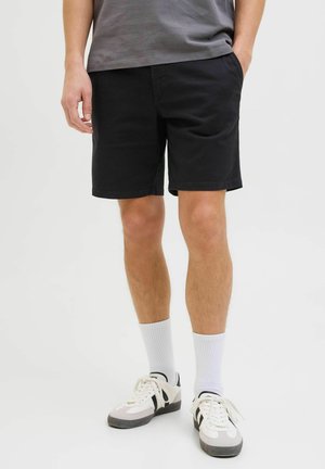Mann trägt schwarze Shorts, weiße Kniestrümpfe und weiße Sneakers mit schwarzen Streifen, steht mit den Händen in den Taschen vor einem einfarbigen Hintergrund.