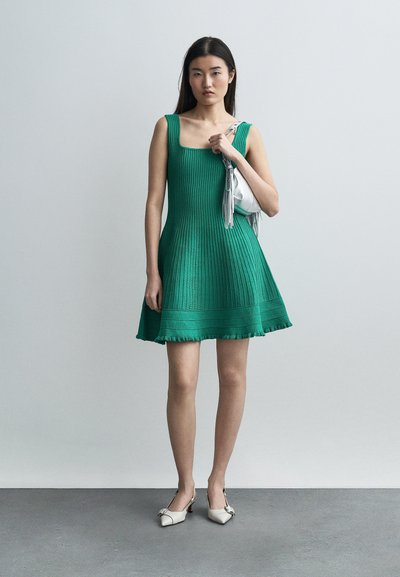 Femme portant une robe verte côtelée sans manches et des talons blancs, tenant un petit sac à main argenté sur l'épaule, sur un fond uni.