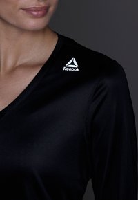 Černý sportovní top s výstřihem do V, na levém rameni s bílým logem Reebok. Materiál působí hladce a lehce.